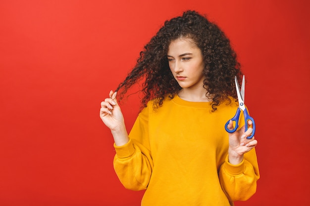 Portrait de jeune fille bouclée choquée coupant ses cheveux avec des ciseaux, isolé sur fond rouge.