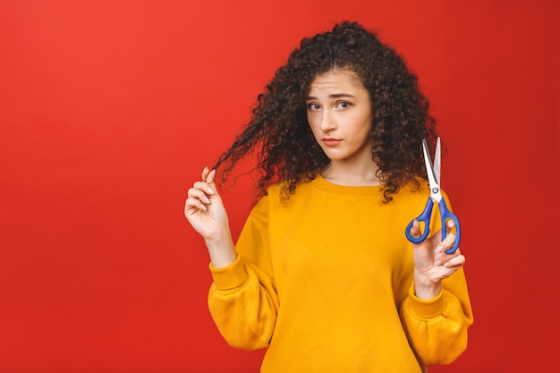 Portrait de jeune fille bouclée choquée coupant ses cheveux avec des ciseaux, isolé sur fond rouge.