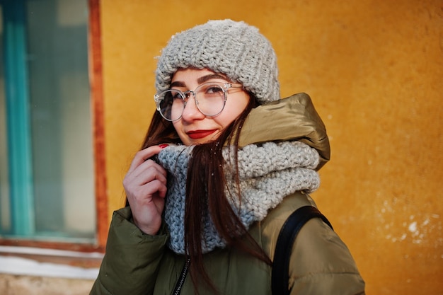 Portrait de jeune fille brune en écharpe grise et lunettes de chapeau par temps froid avec soleil contre le mur orange de la vieille maison