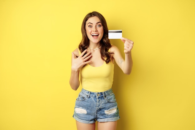 Portrait de jeune fille brune montrant sa carte de crédit et souriant, portant des vêtements d'été, concept de banque, argent et shopping, debout sur fond jaune