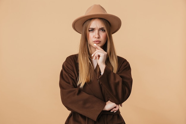 Portrait de jeune fille caucasienne portant un chapeau et un manteau pensant avec un regard sombre isolé sur beige