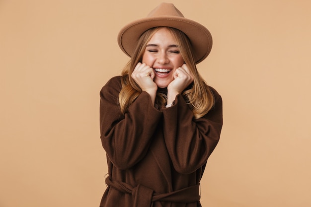 Portrait de jeune fille caucasienne portant chapeau et manteau riant isolé sur beige