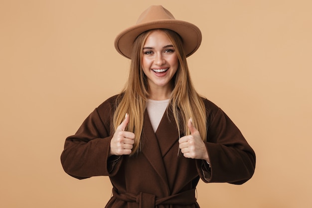 Portrait de jeune fille caucasienne portant chapeau et manteau souriant et montrant les pouces vers le haut isolé sur beige