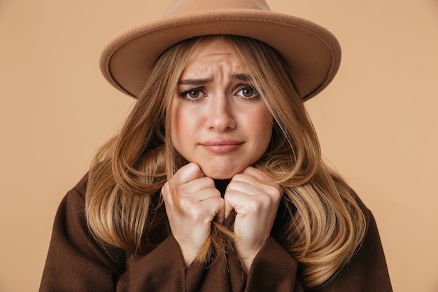 Portrait de jeune fille caucasienne portant un chapeau et un manteau tremblant et se sentant froid isolé sur beige