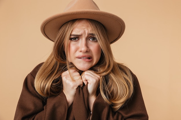 Portrait de jeune fille caucasienne portant un chapeau et un manteau tremblant et se sentant froid isolé sur beige