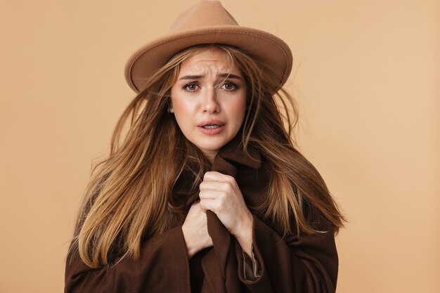 Portrait de jeune fille caucasienne portant un chapeau et un manteau tremblant et se sentant froid isolé sur beige