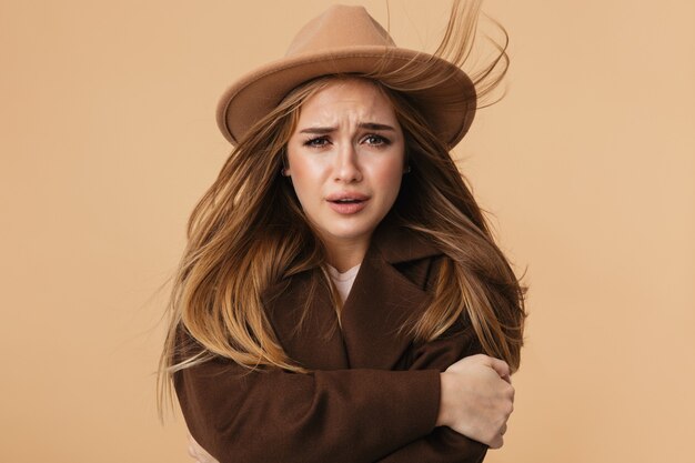 Portrait de jeune fille caucasienne portant un chapeau et un manteau tremblant et se sentant froid isolé sur beige