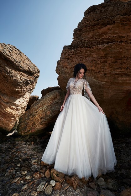 Portrait d'une jeune fille dans une robe magnifique de mariage posant un photographe sur la plage. La mariée est sur les rochers