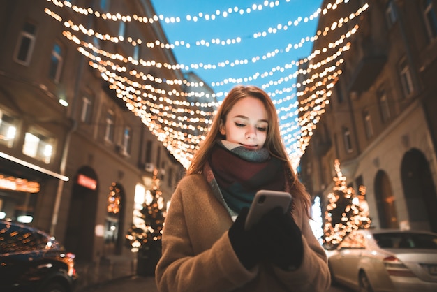 Portrait d'une jeune fille dans des vêtements chauds debout à l'extérieur sur un fond de lumières bokeh et utiliser un smartphone