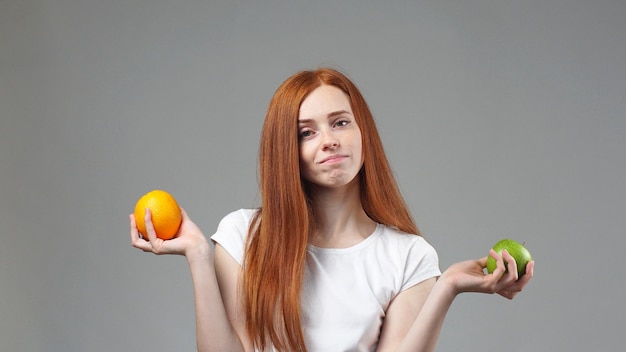 Portrait d'une jeune fille debout sur un mur gris s'est demandé quel fruit est plus sain Apple ou orange