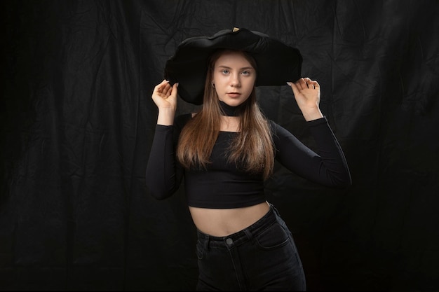 Portrait de jeune fille élégante en haut moulant court et chapeau à larges bords en studio sur fond noir Jeune sorcière Halloween