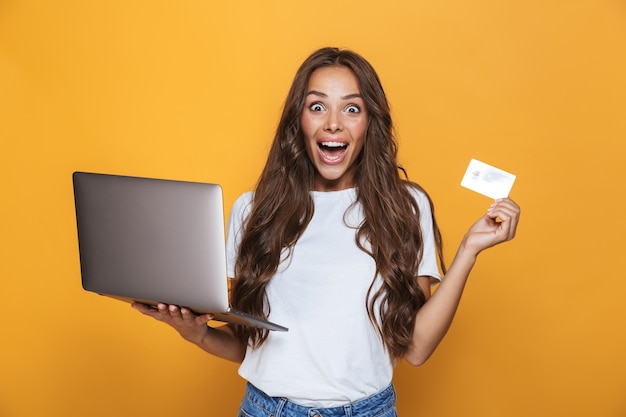 Portrait d'une jeune fille excitée avec de longs cheveux brune debout sur un mur jaune, tenant un ordinateur portable, montrant une carte de crédit en plastique