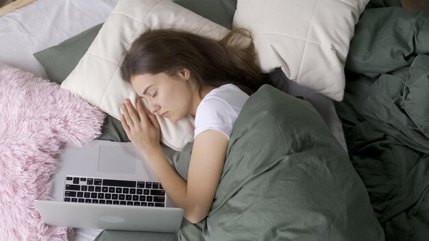 Portrait d'une jeune fille fatiguée endormie avec un ordinateur portable au lit à la maison bureau à domicile