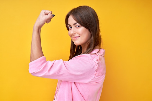 Portrait de jeune fille féministe brune montre la force, démontre les biceps et regarde la caméra en jaune. Concept de puissance féminine