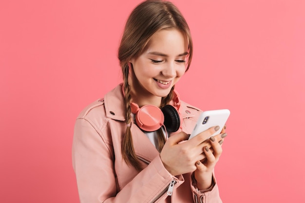 Portrait de jeune fille gaie avec deux tresses en veste de cuir avec un casque utilisant joyeusement un téléphone portable sur fond rose isolé