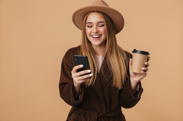 Portrait de jeune fille heureuse portant un chapeau tenant un téléphone portable et buvant du café isolé sur beige