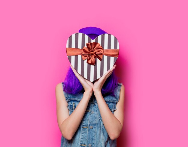 Portrait de jeune fille hipster de style avec une coiffure violette avec boîte-cadeau Saint Valentin sur fond rose