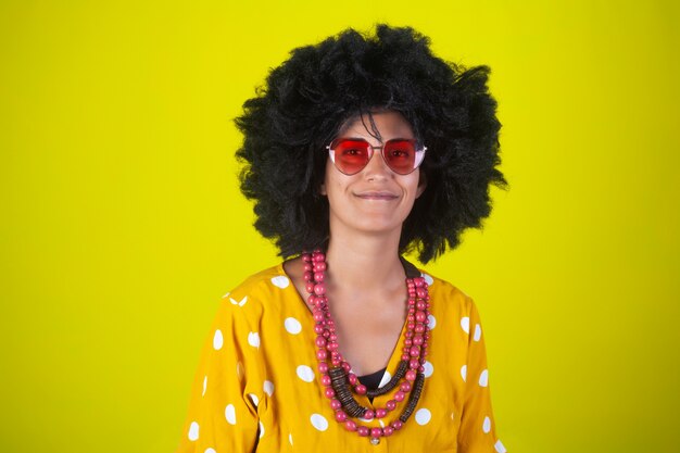 Portrait d'une jeune fille indienne souriante avec coiffure afro bouclée et lunettes en forme de coeur sur mur jaune