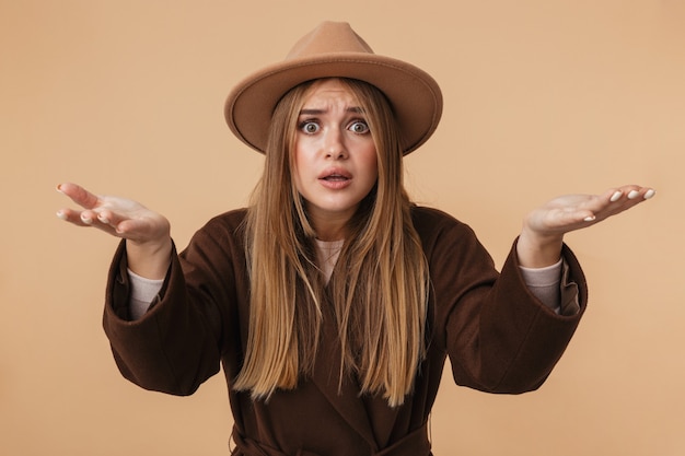 Portrait de jeune fille inquiète portant un chapeau et un manteau tendant les bras isolés sur beige