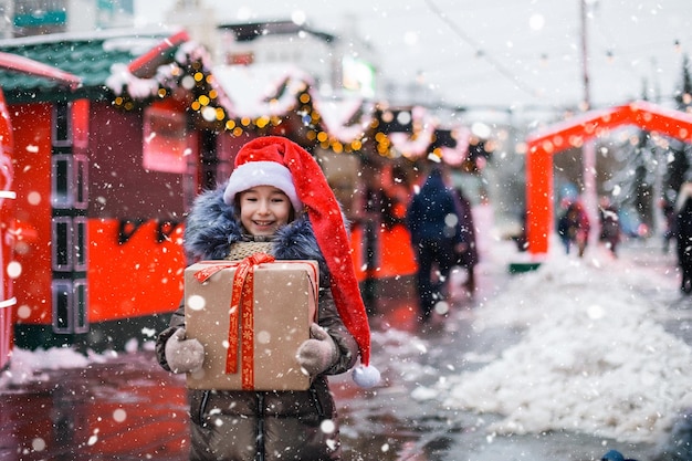 Portrait de jeune fille joyeuse en bonnet de Noel avec boîte-cadeau pour Noël dans la rue de la ville en hiver avec de la neige sur le marché festif avec des décorations et des guirlandes Vêtements chauds écharpe tricotée et fourrure Nouvel an