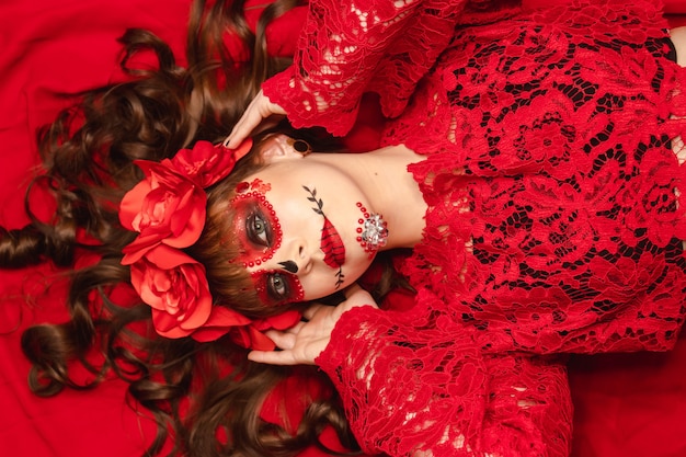 Portrait d'une jeune fille maquillée et tenue Dia de los Muertos allongée sur fond rouge.