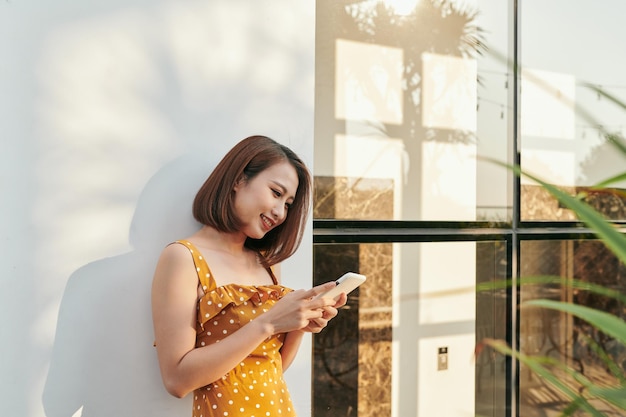 Portrait de jeune fille de mode souriante debout le mur et à l'aide de smartphone