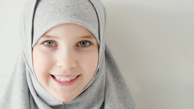 Portrait d'une jeune fille musulmane séduisante de 9 ans portant un hijab gris et une robe traditionnelle regardant la caméra et souriant sur fond clair, espace de copie.