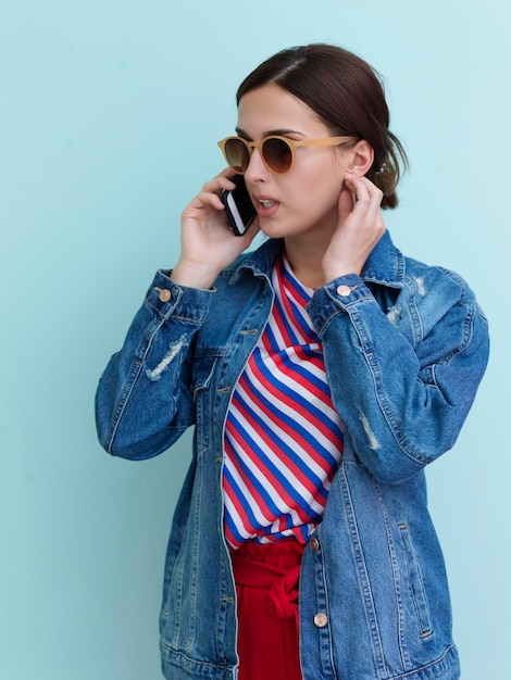 Portrait de jeune fille parlant au téléphone en se tenant debout devant un fond bleu. Modèle féminin portant des lunettes de soleil représentant le concept de mode et de technologie moderne