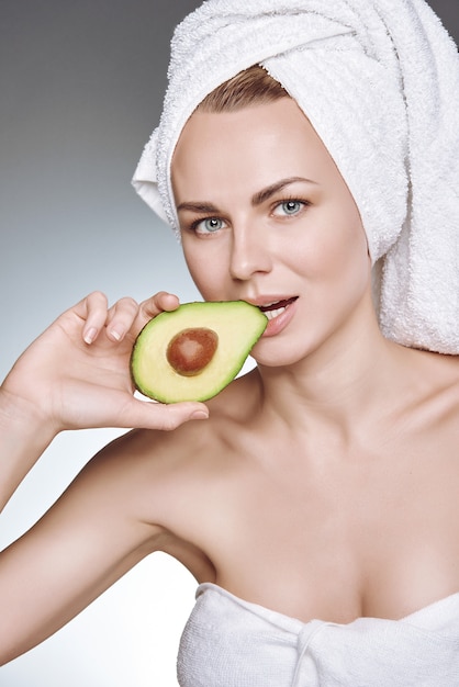 Portrait d'une jeune fille à la peau saine et soyeuse, avec une serviette blanche sur la tête tenant une tranche d'avocat avec une pierre