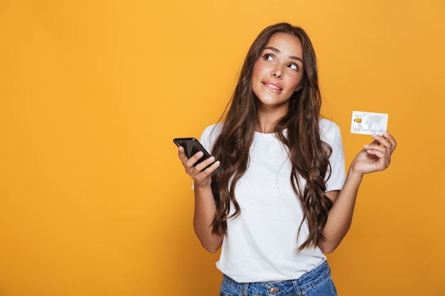 Portrait d'une jeune fille pensive avec de longs cheveux brune debout sur un mur jaune, tenant un téléphone mobile, montrant une carte de crédit en plastique