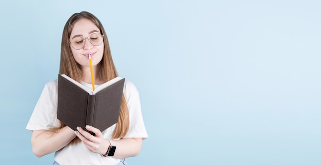 Portrait de jeune fille pensive réfléchie ayant un cahier et un crayon dans la bouche. Isolé sur fond bleu avec espace copie.