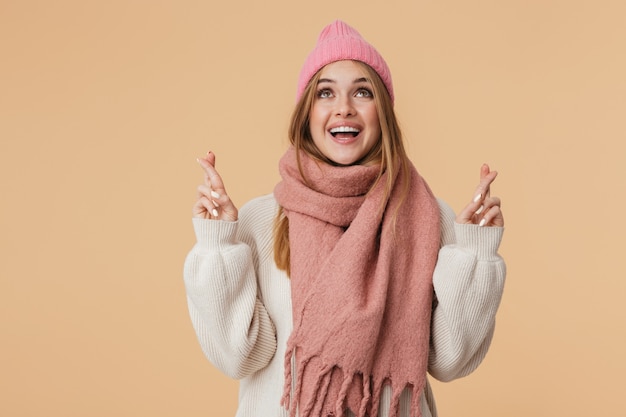 Portrait de jeune fille portant un chapeau d'hiver et une écharpe souriant et gardant les doigts croisés isolés sur beige