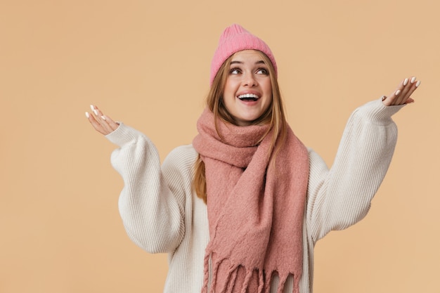 Portrait de jeune fille portant un chapeau d'hiver et une écharpe souriant et regardant vers le haut avec les bras grands ouverts isolé sur beige