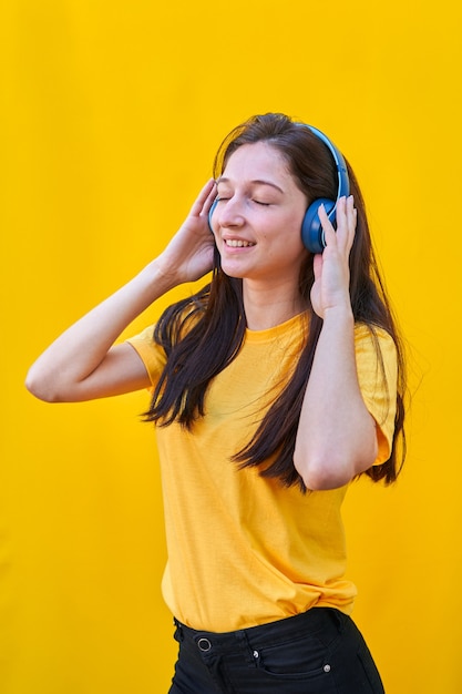 Portrait d'une jeune fille de race blanche aux longs cheveux bruns, t-shirt jaune et jean noir, écoutant de la musique avec ses écouteurs bleus.