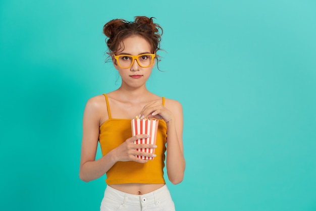 Portrait d'une jeune fille en riant dans des vêtements décontractés tenant une boîte de pop-corn et regardant la caméra isolée sur lumineux