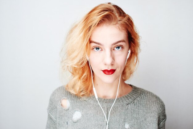 Portrait d'une jeune fille rousse sur fond gris dans un pull écoutant de la musique avec des écouteurs