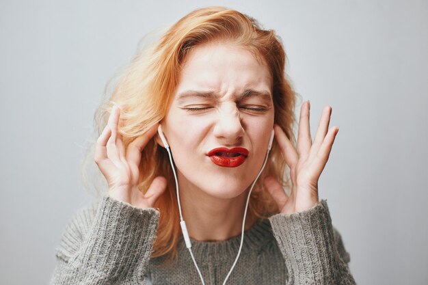 Portrait d'une jeune fille rousse sur fond gris dans un pull écoutant de la musique avec des écouteurs
