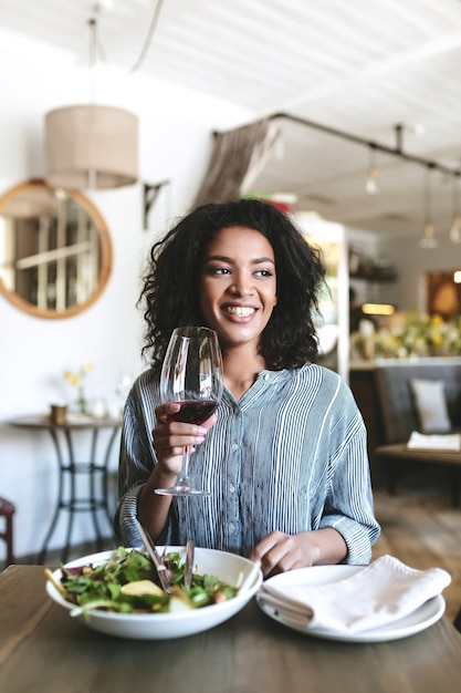 Portrait de jeune fille souriante aux cheveux bouclés sombres assis au restaurant avec verre de vin rouge et salade sur table