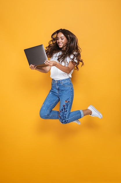 Portrait d'une jeune fille souriante aux longs cheveux noirs sautant par-dessus le mur jaune, à l'aide d'un ordinateur portable