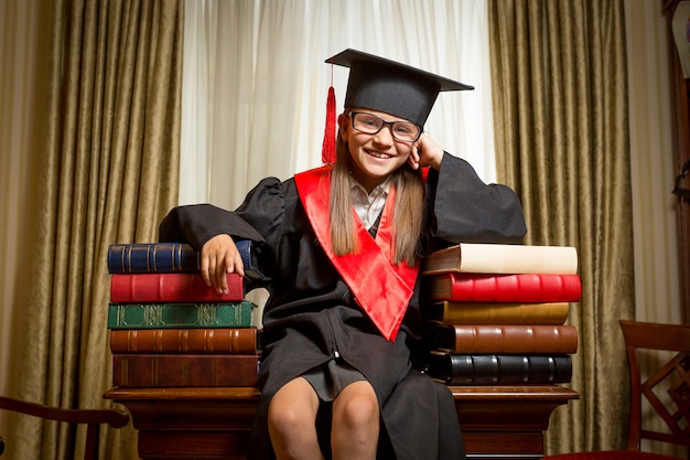 Portrait de jeune fille souriante en chapeau de graduation et robe assise sur une table et s'appuyant sur des livres