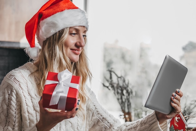 Portrait d'une jeune fille souriante dans un chapeau de Noël festif tenant une boîte-cadeau rouge et appelant par vidéoconférence