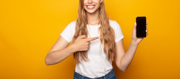 Portrait de jeune fille souriante pointant avec le doigt sur smartphone isolé sur jaune