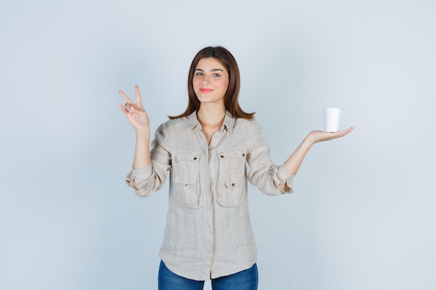 Portrait de jeune fille tenant une tasse de café en plastique, montrant le signe V en chemise et l'air gai