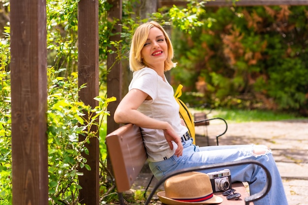 Portrait d'une jeune fille touristique blonde en vacances d'été assis sur un banc dans un parc portant un sac à dos jaune appareil photo chapeau