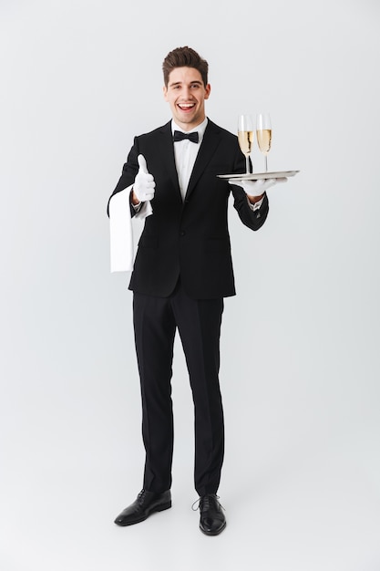 Portrait d'un jeune garçon souriant en smoking tenant un plateau avec deux verres de champagne sur un mur blanc