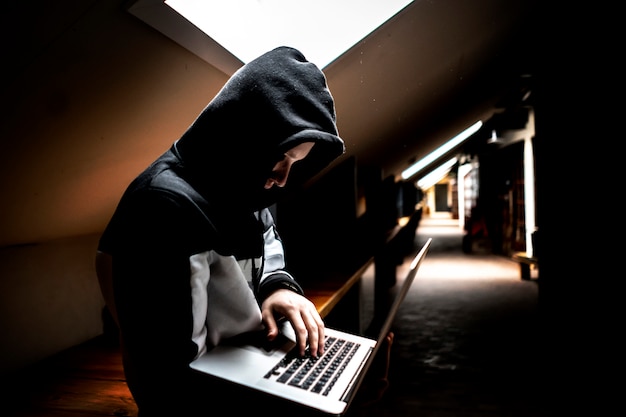 Portrait de jeune geek en cagoule dans une pièce sombre avec un ordinateur portable