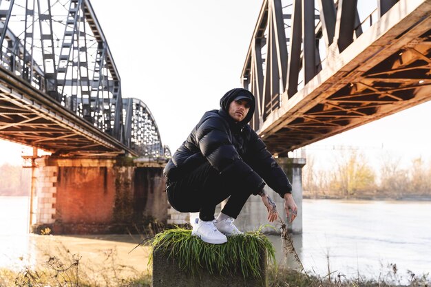 Photo portrait d'un jeune homme accroupi sur la terre près des ponts de la ville