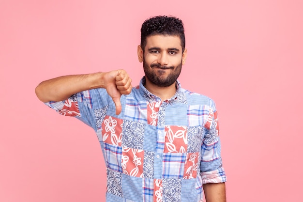 Portrait d'un jeune homme adulte en chemise bleue critiquant la mauvaise qualité avec les pouces vers le bas grimace mécontente, montrant un geste d'aversion, exprimant sa désapprobation. Tourné en studio intérieur isolé sur fond rose.