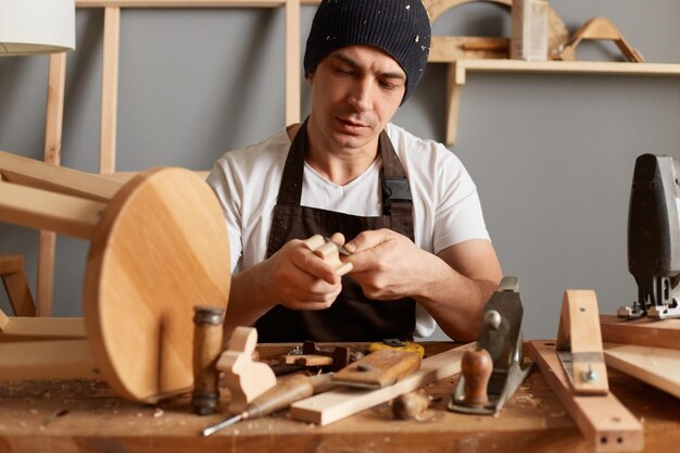 Portrait d'un jeune homme adulte concentré charpentier portant un t-shirt blanc, une casquette noire et un tablier marron travaillant des travaux de menuiserie des ciseaux à bois pour la sculpture sur le bureau du menuisier des travaux de menuiserie en bois