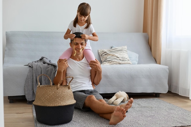 Portrait d'un jeune homme adulte positif portant un t-shirt blanc de style décontracté assis sur le sol près du canapé avec sa fille, petite fille assise sur les épaules du père et lui faisant une coiffure.